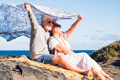 Happy Senior Couple Sitting on Beach holding Blanket In Wind What is an IUL San Diego CA